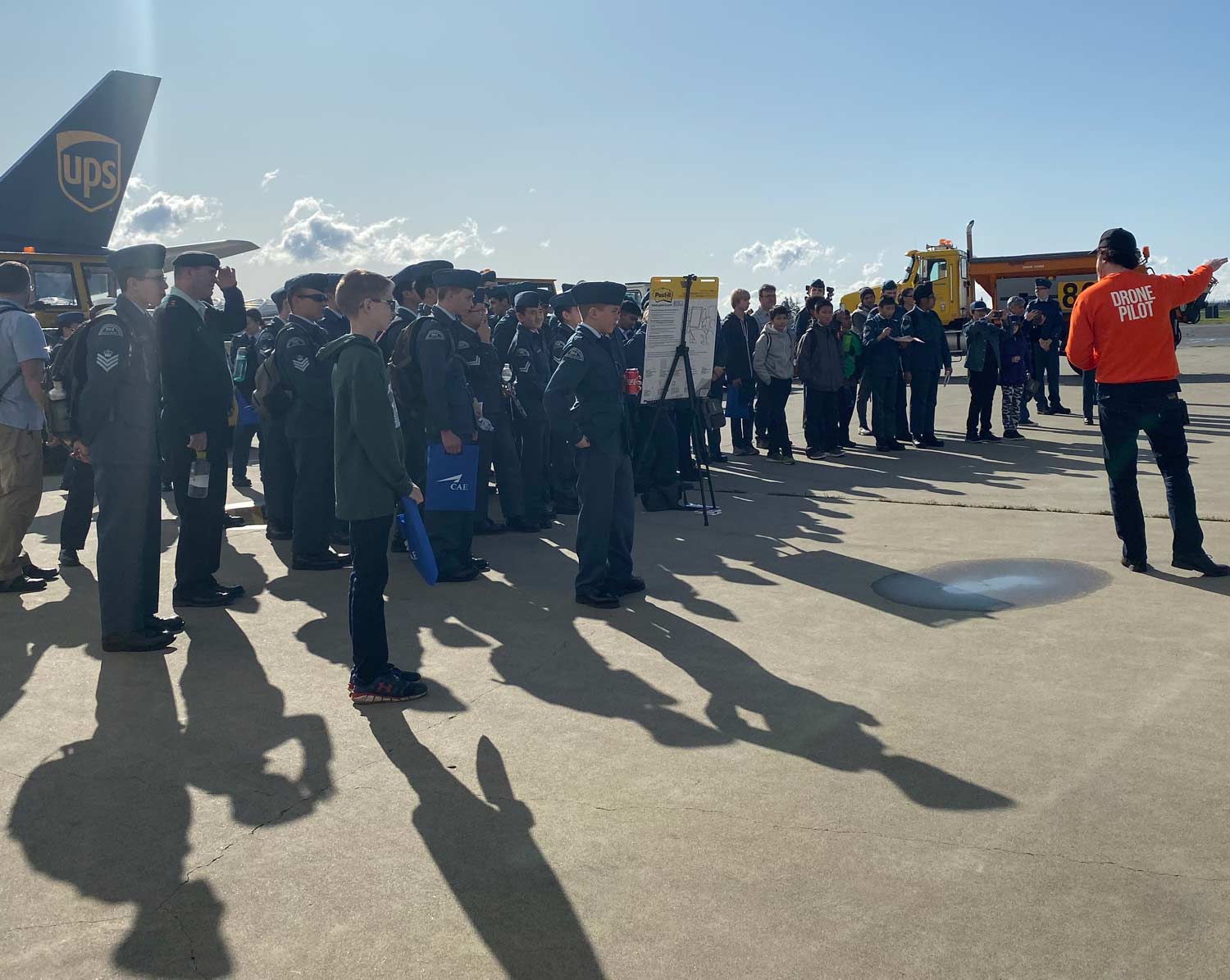 11 Alpha drone pilot in an orange flight shirt briefing cadets on an airport apron